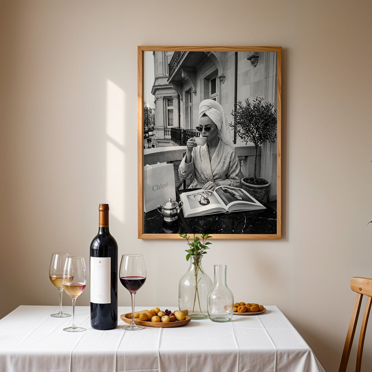 Woman in towel turban drinking coffee on balcony print in an oak frame above a table set with red wine and glasses