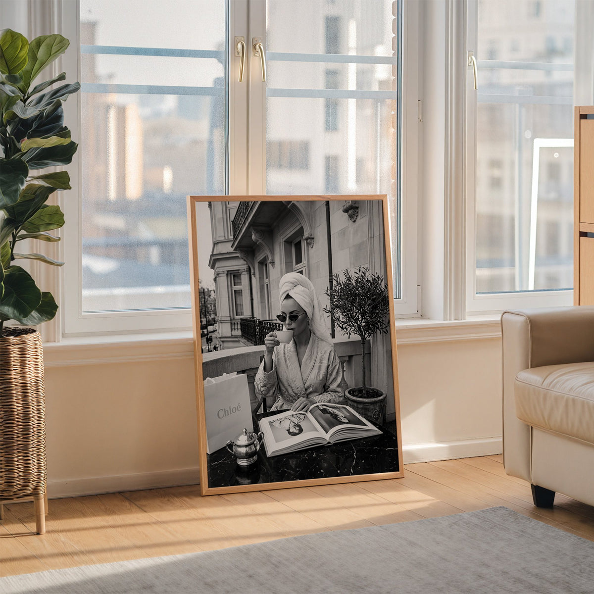 Woman in towel turban drinking coffee on balcony print in an oak frame leaning on the floor beside a fiddle leaf fig