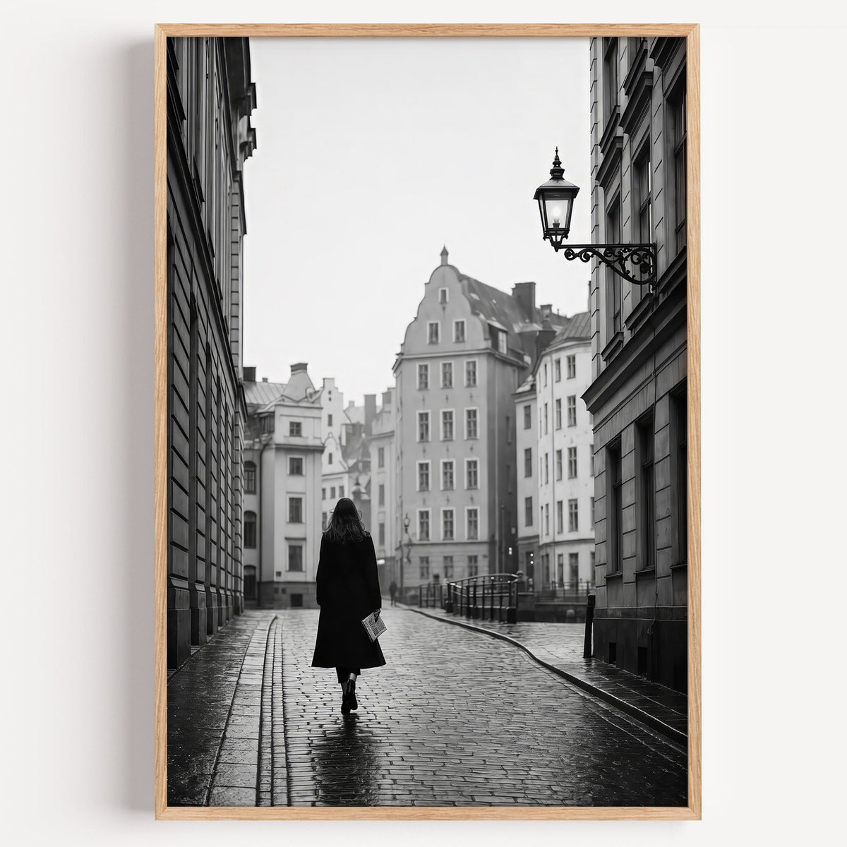 Woman walking on cobblestone old town street print in a light oak frame on a plain white wall