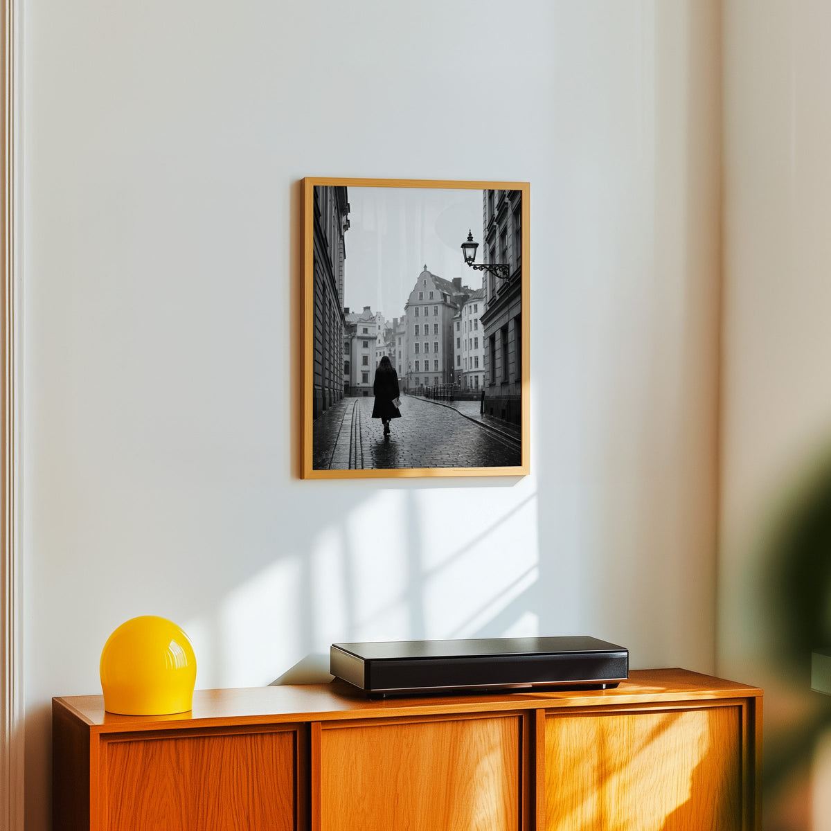Woman walking on cobblestone old town street print in an oak frame above a teak sideboard with a yellow glass lamp