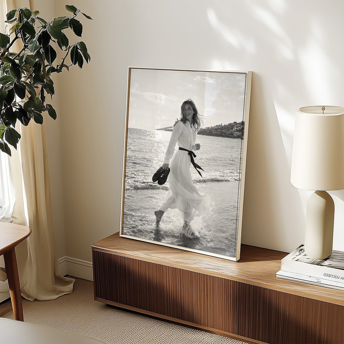 Laughing woman in white dress running in the surf print in a white frame on a walnut sideboard beside a ceramic lamp