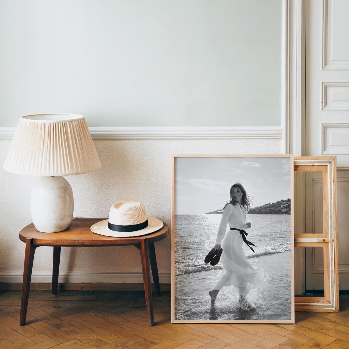 Laughing woman in white dress running in the surf print in a pine frame on a wood floor beside a lamp and straw hat