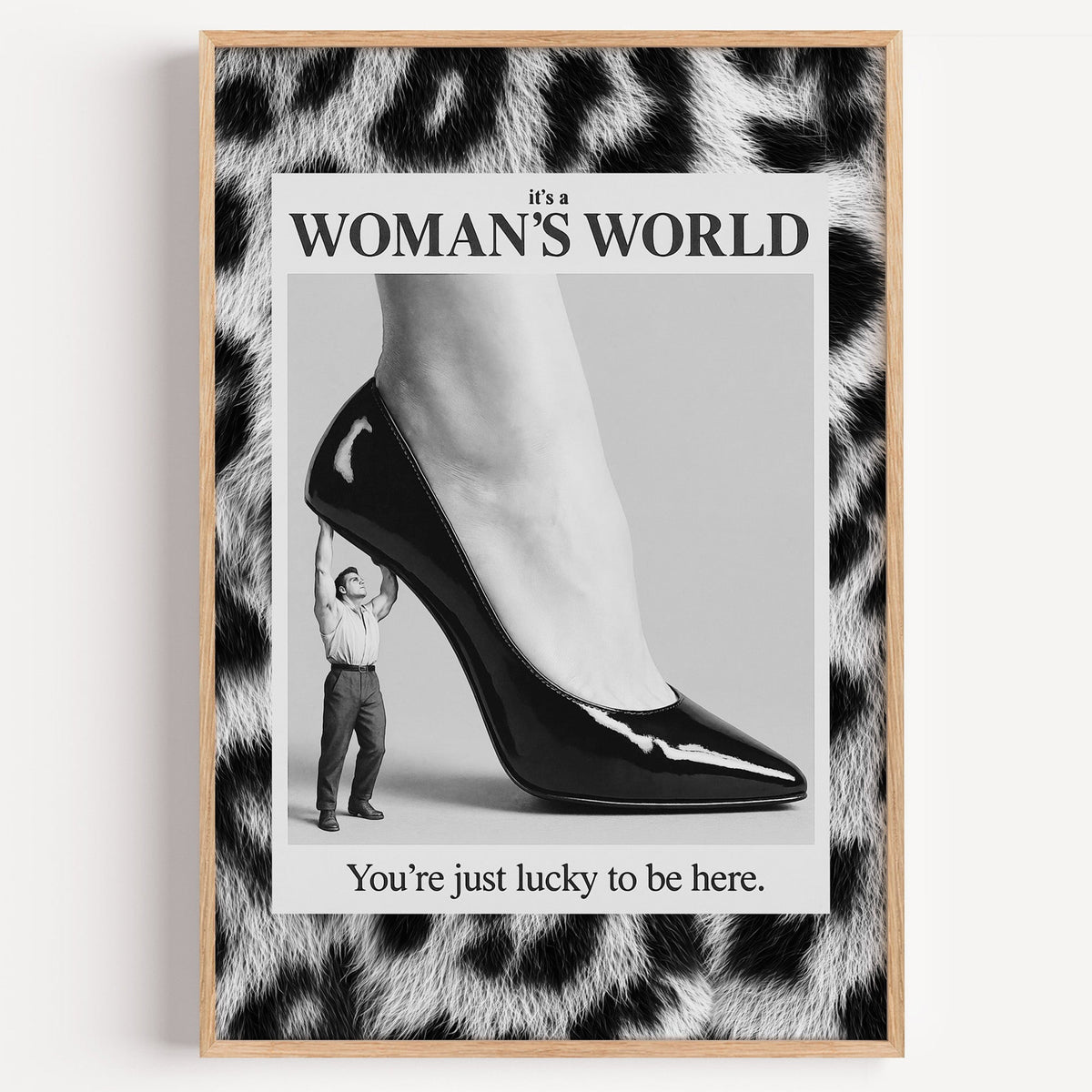 Tiny man lifting giant stiletto in leopard frame print in a light oak frame on a plain white wall