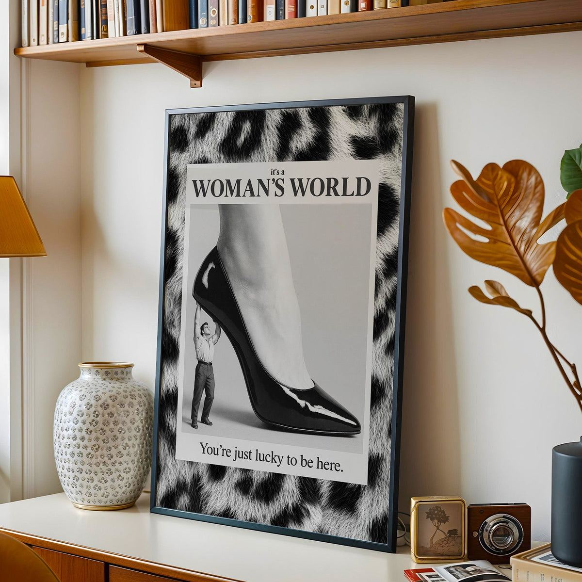 Tiny man lifting giant stiletto in leopard frame print in a black frame on a console under a bookshelf