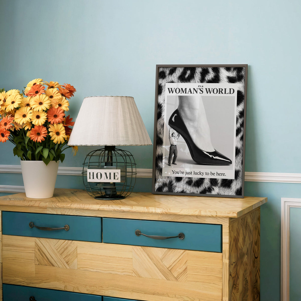 Tiny man lifting giant stiletto in leopard frame print in a charcoal frame on a teal dresser beside orange daisies