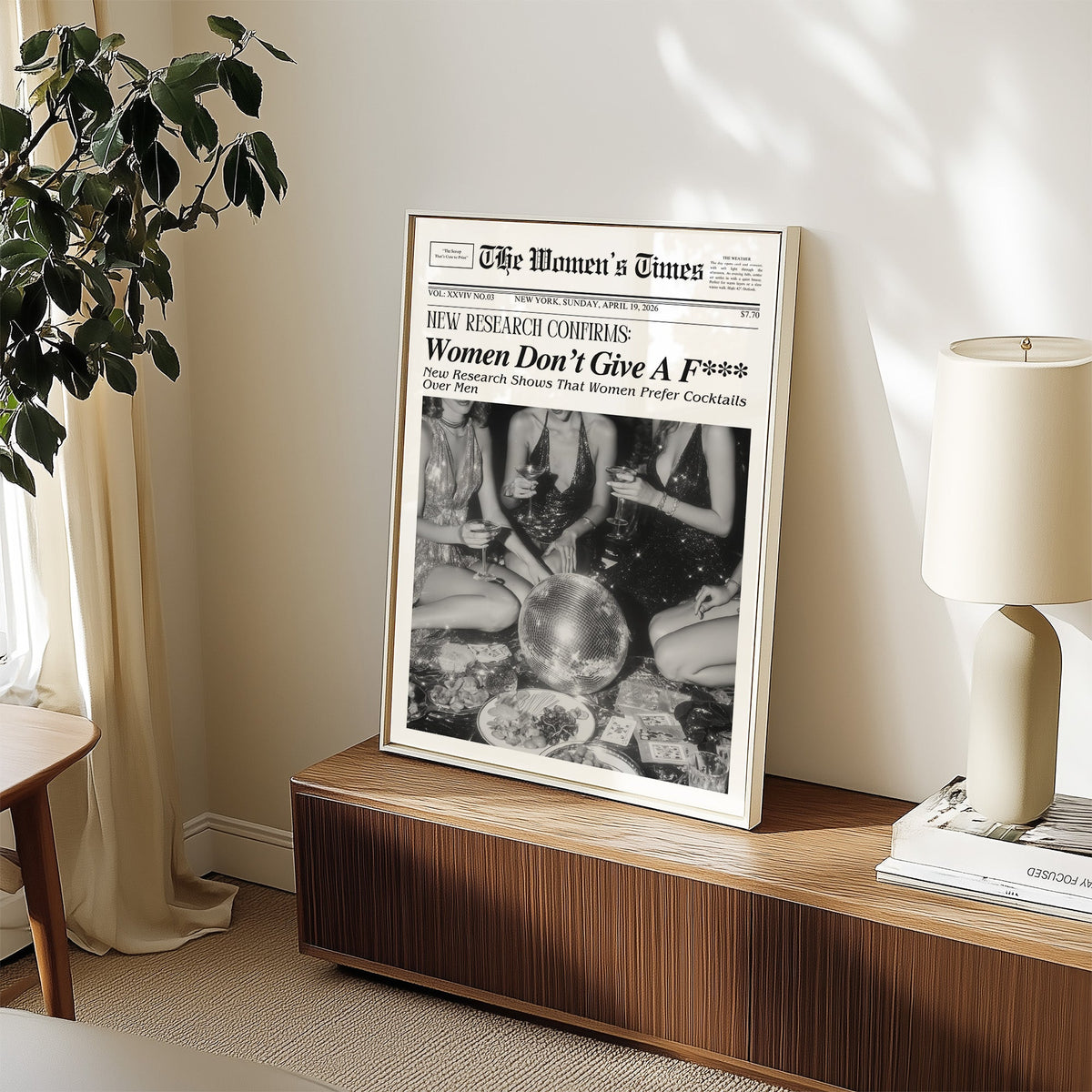 Mock front page with women around a disco ball print in a white frame on a walnut sideboard beside a ceramic lamp