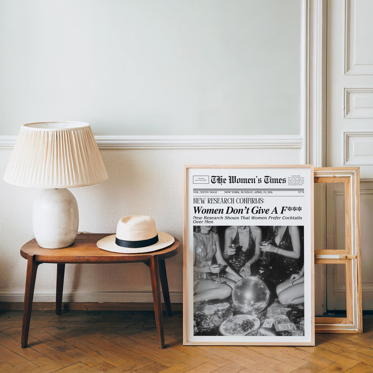 Mock front page with women around a disco ball print in a pine frame on a wood floor beside a lamp and straw hat