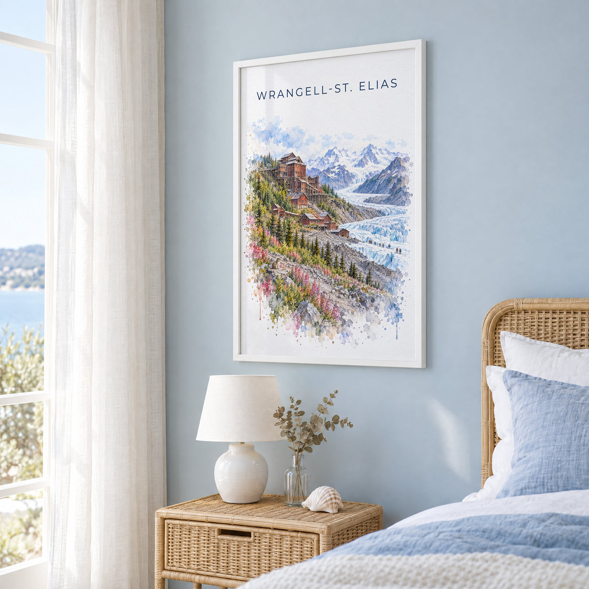 Wrangell-St. Elias National Park watercolor in a white frame on a pale blue wall beside a rattan bed in a coastal bedroom