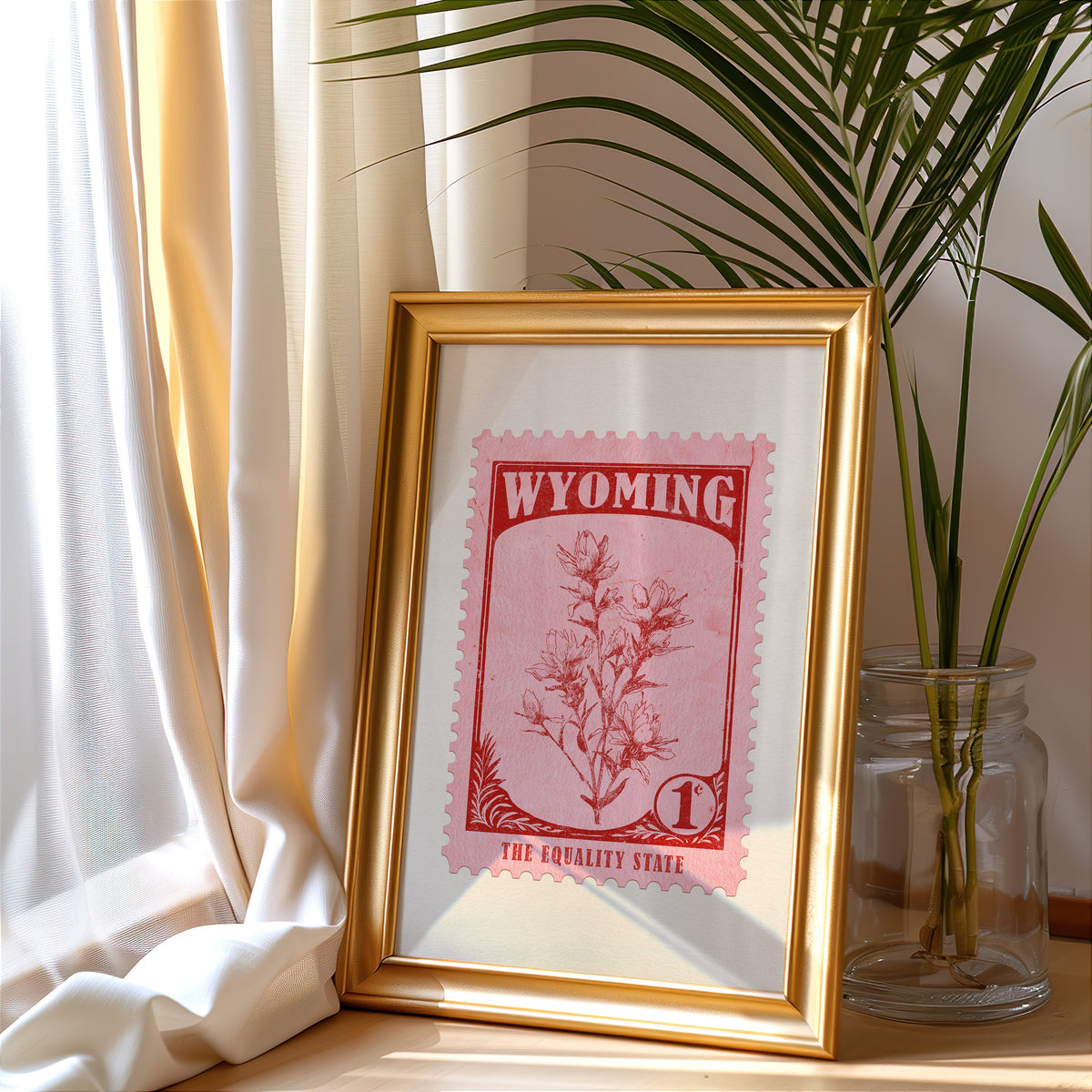 Wyoming stamp print in a gold frame leaning by a cream curtain with a palm in a glass jar