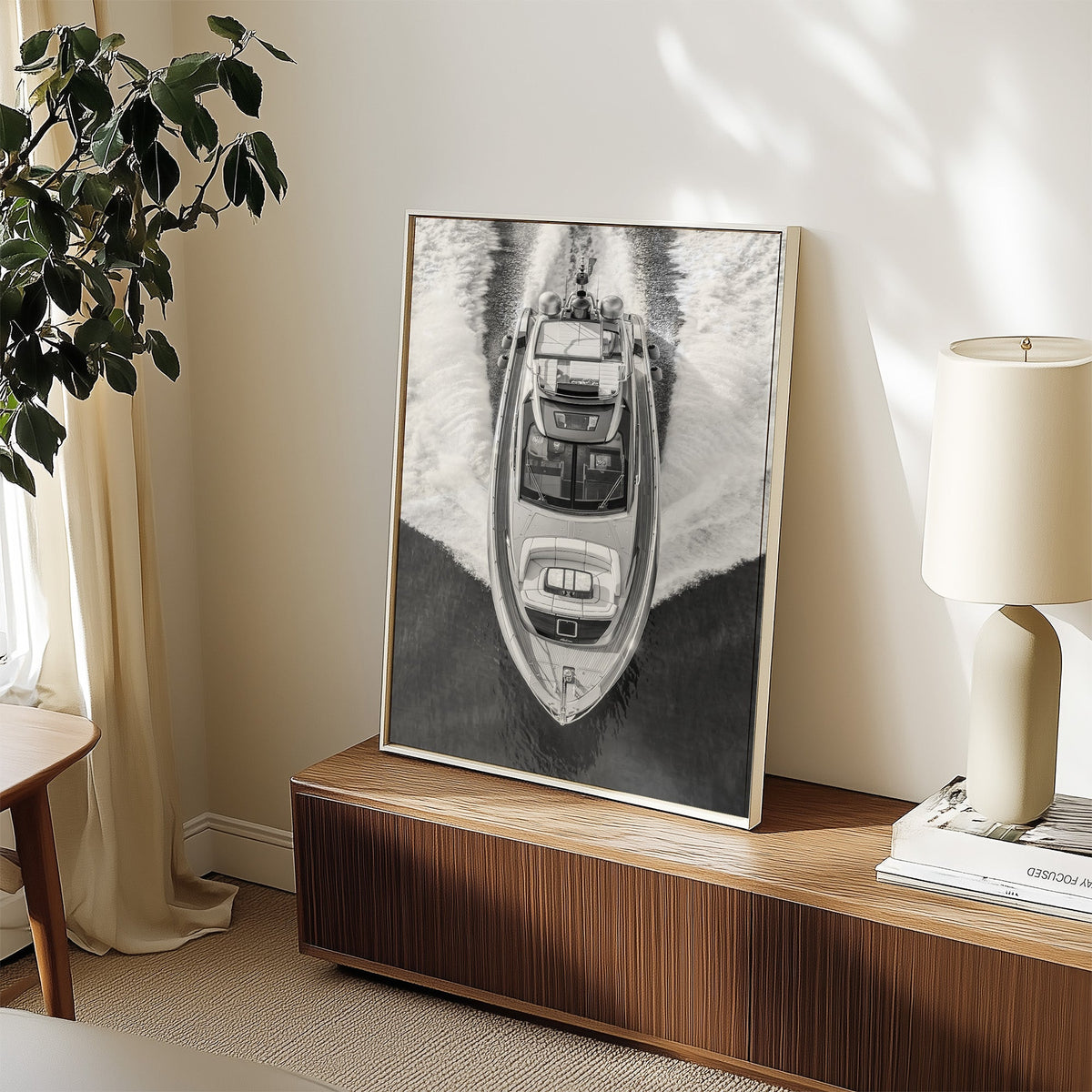 Aerial view of a yacht with white wake print in a white frame on a walnut sideboard beside a ceramic lamp