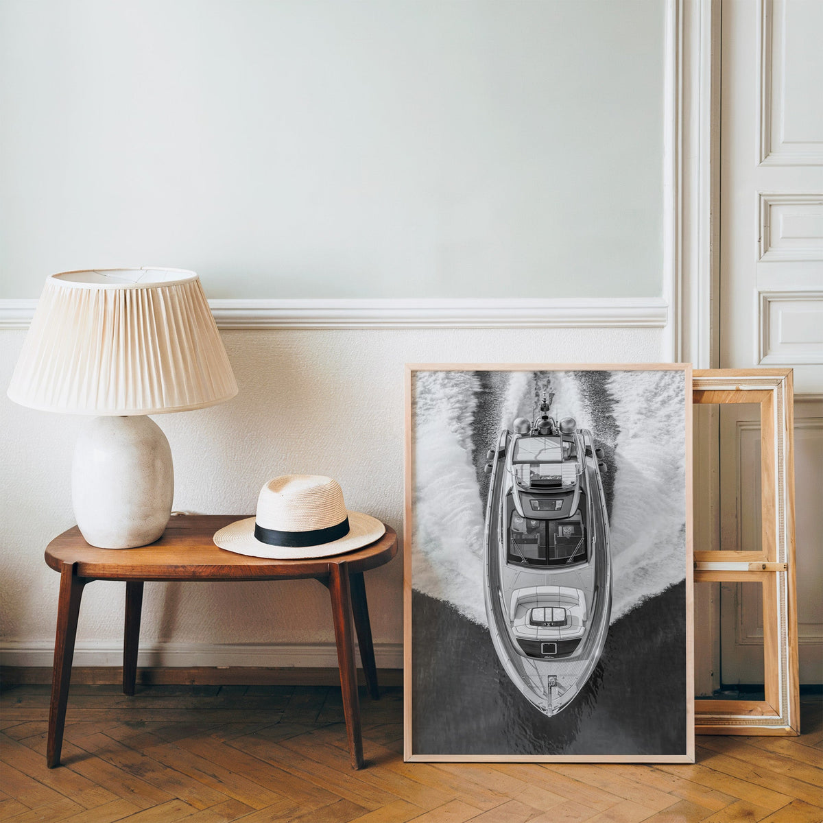 Aerial view of a yacht with white wake print in a pine frame on a wood floor beside a lamp and straw hat