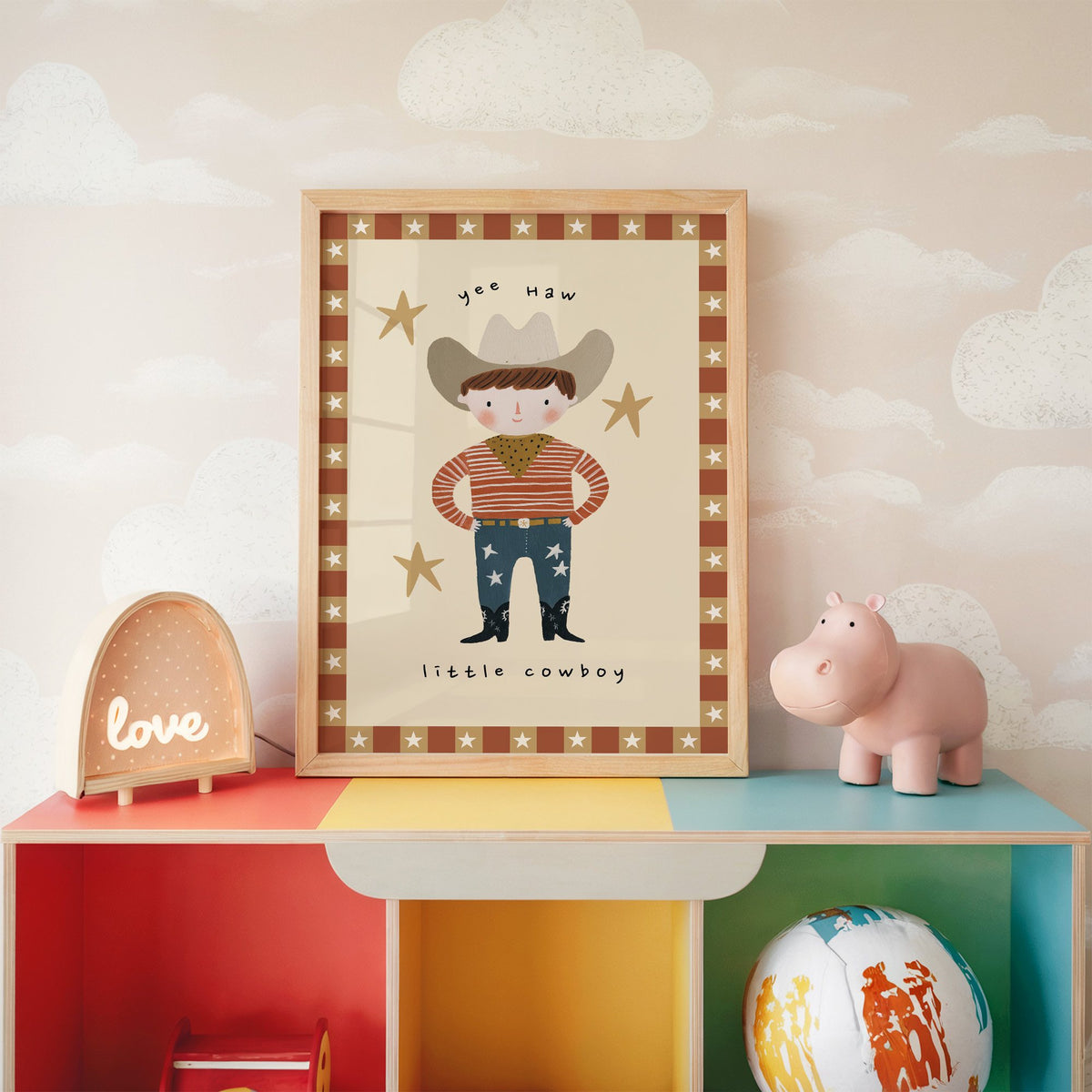 Yee haw cowboy star border nursery print in an oak frame on a rainbow toy shelf against cloud wallpaper