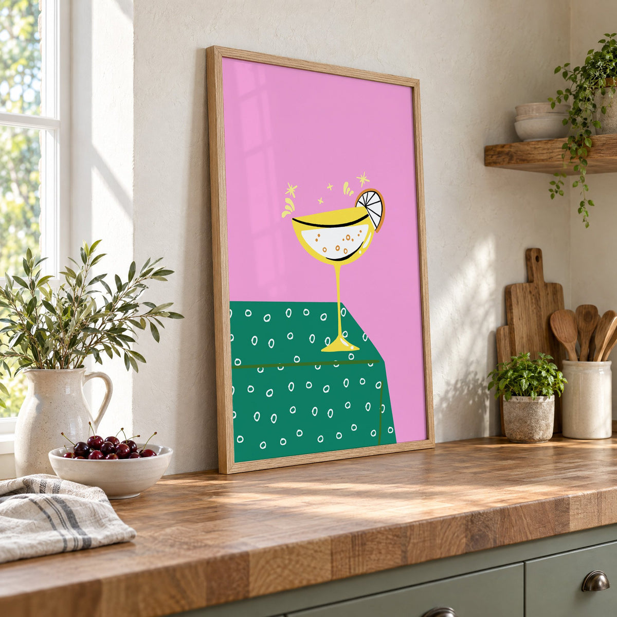 Yellow coupe cocktail print in an oak frame leaning on a butcher block island beside a bowl of cherries