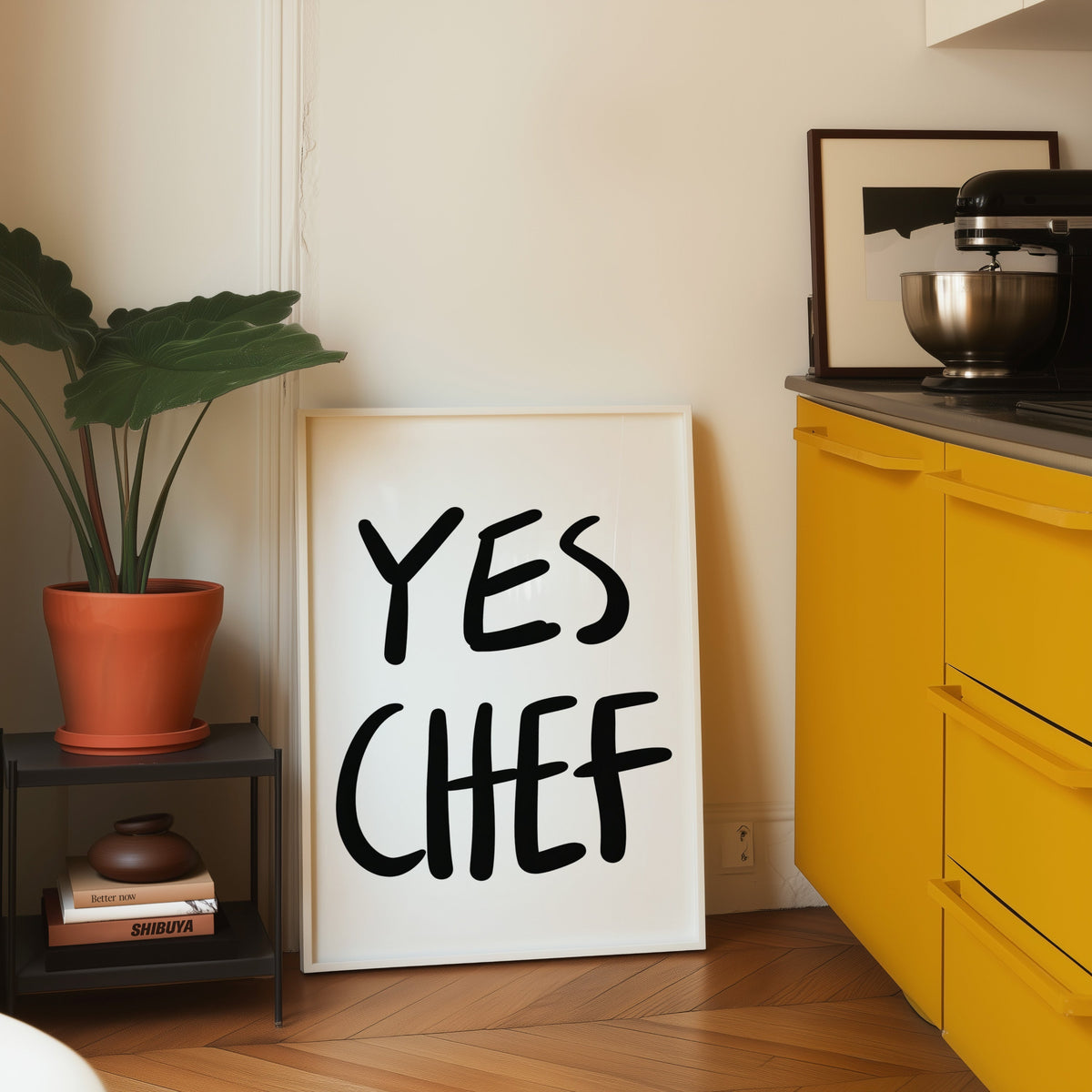 Yes Chef print in a white frame leaning on parquet beside a bright yellow kitchen