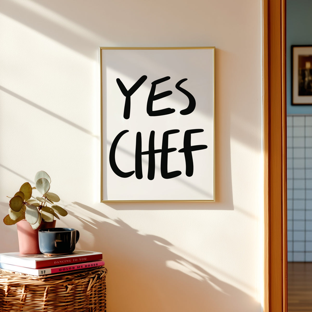Yes Chef print in a gold frame above a basket of books and a pilea plant