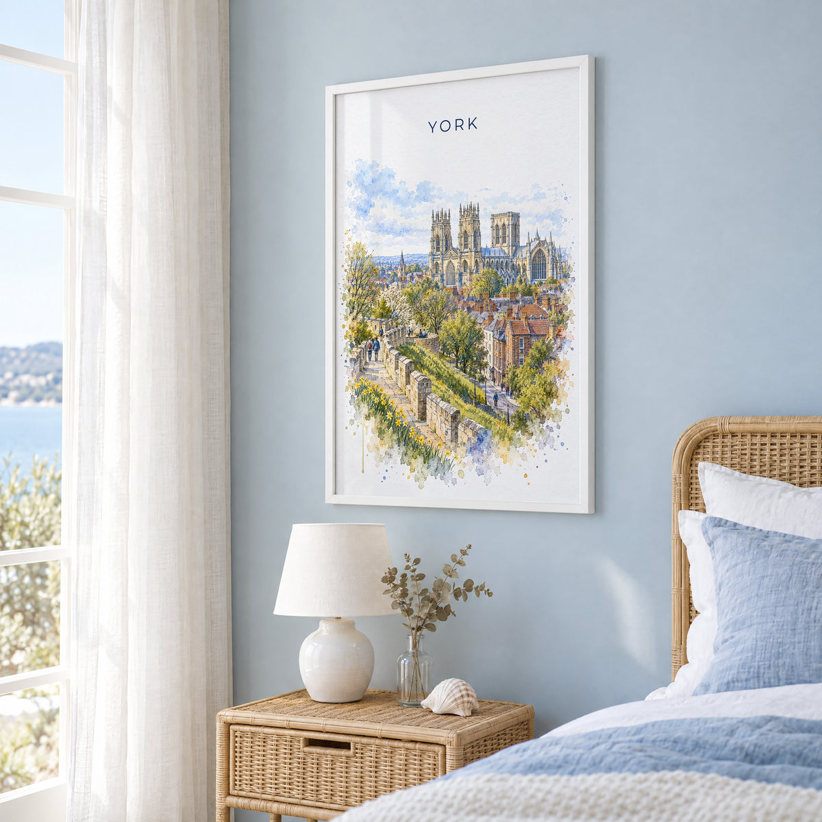 York, England watercolor print in a white frame on a pale blue wall beside a rattan bed in a coastal bedroom