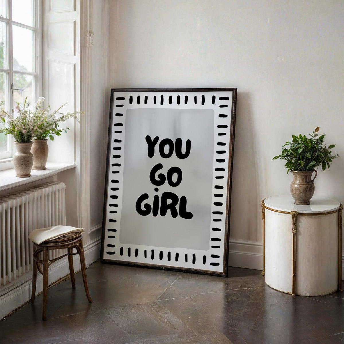 You go girl bubble letter quote print in a black frame on a dark stone floor beside a white side table