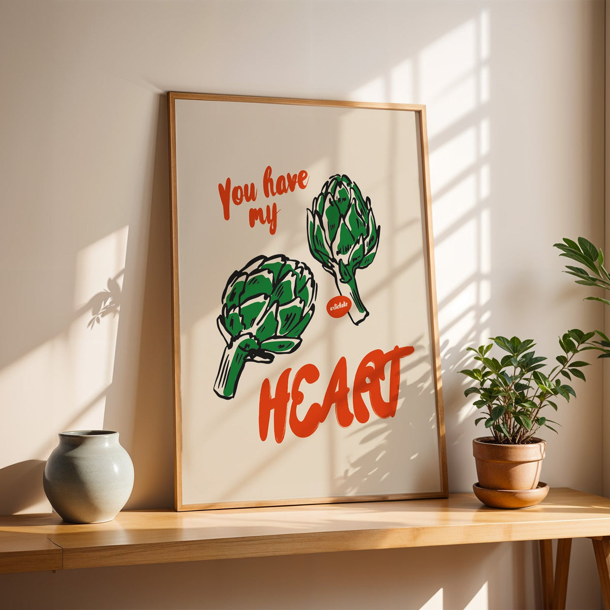Artichoke print in a slim oak frame on a console with a grey-blue vase and leaf shadows