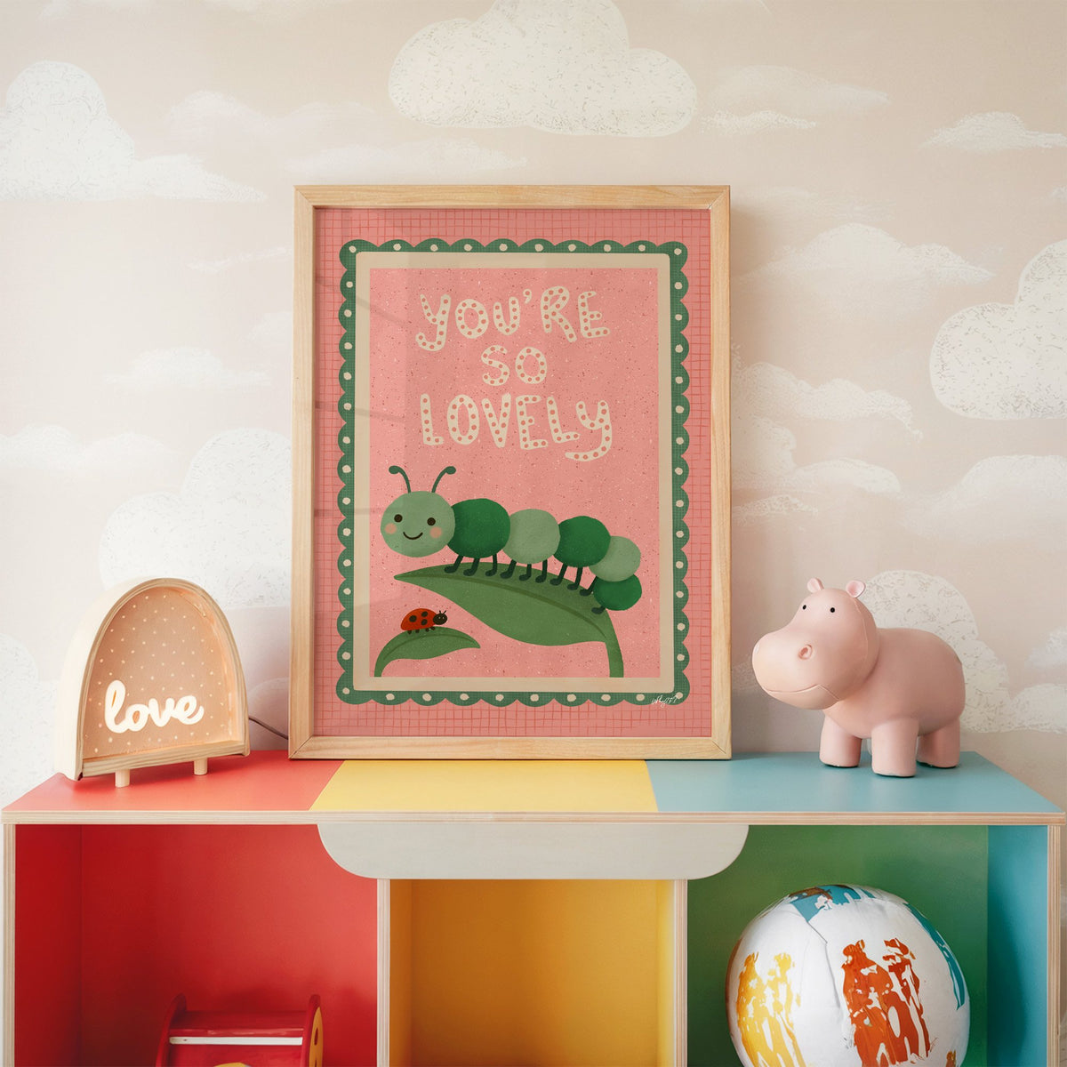 Smiling caterpillar on a leaf nursery print in an oak frame on a rainbow toy shelf against cloud wallpaper