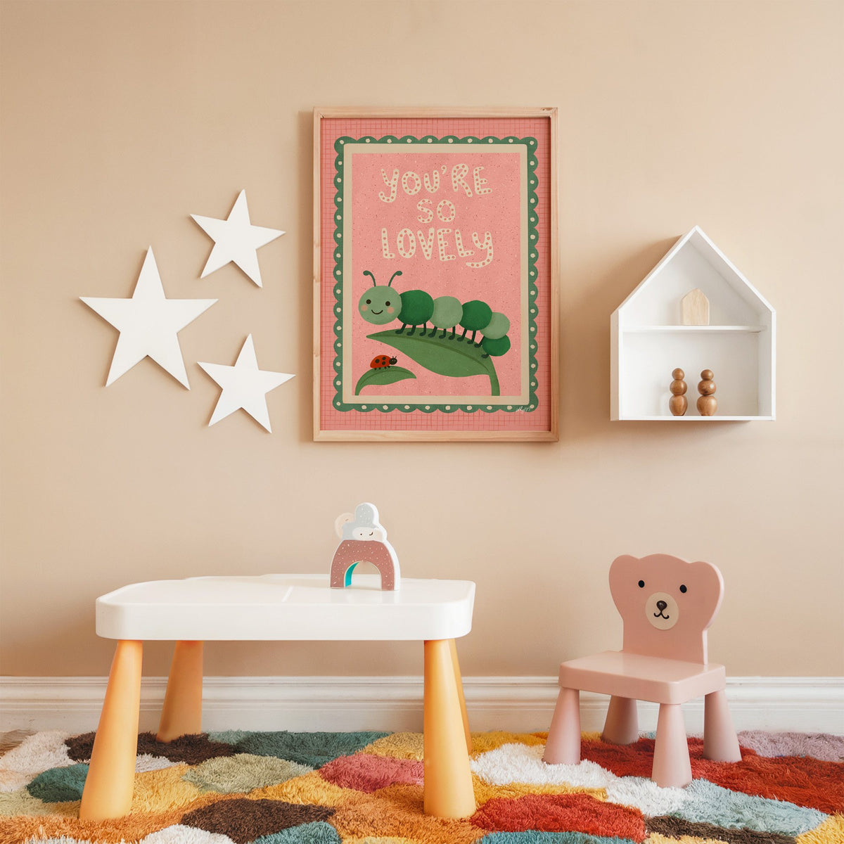Smiling caterpillar on a leaf nursery print in an oak frame on a peach wall above a kids table with wall stars