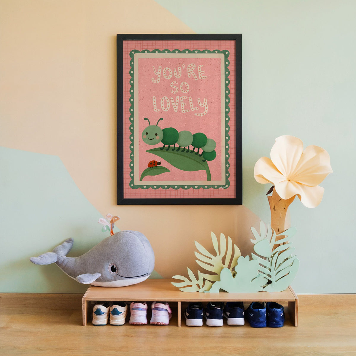Smiling caterpillar on a leaf nursery print in a black frame on a mint and peach wall above a whale plush