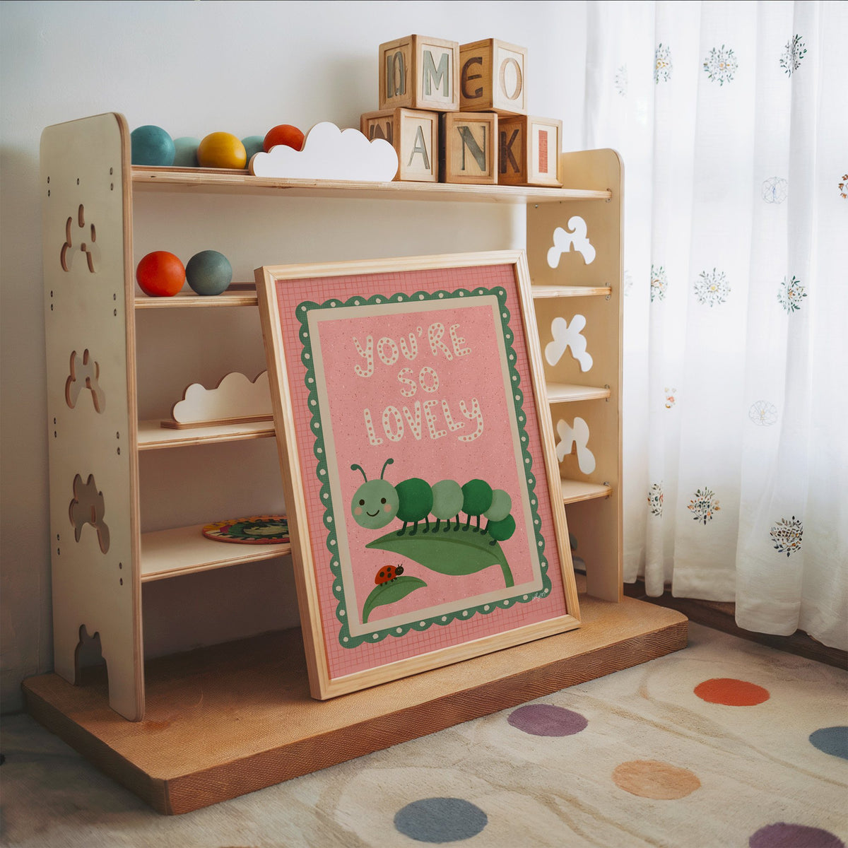 Smiling caterpillar on a leaf nursery print in an oak frame leaning on a toy shelf with wooden letter blocks