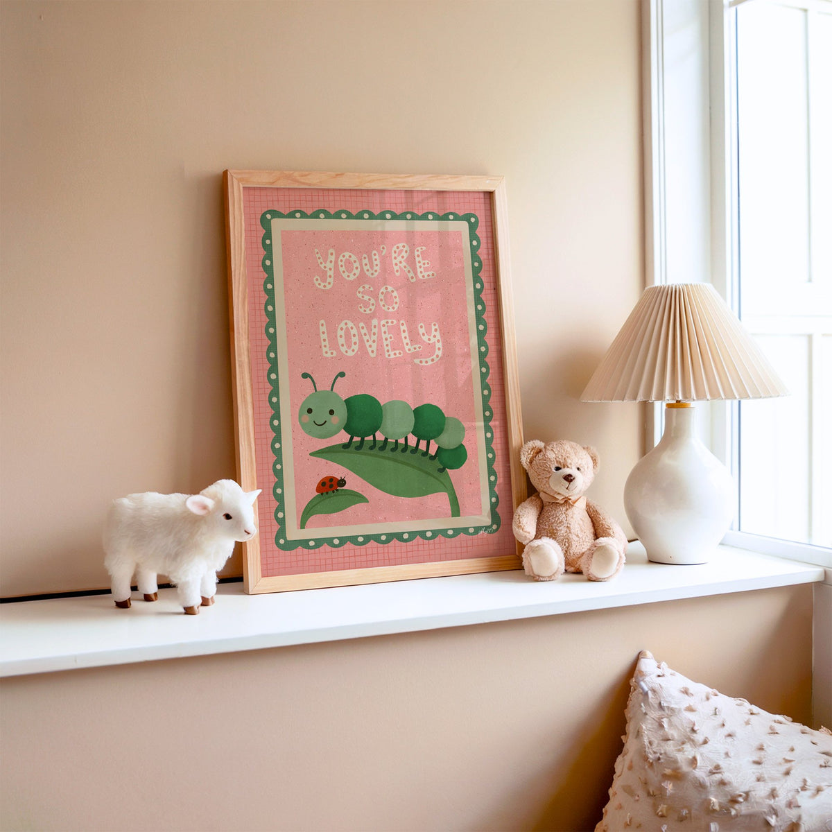 Smiling caterpillar on a leaf nursery print in an oak frame on a window ledge with a toy lamb and teddy