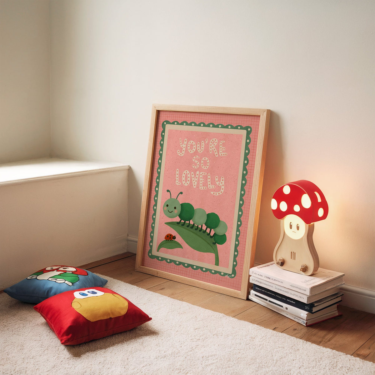 Smiling caterpillar on a leaf nursery print in an oak frame leaning on a rug beside a mushroom lamp and floor cushions